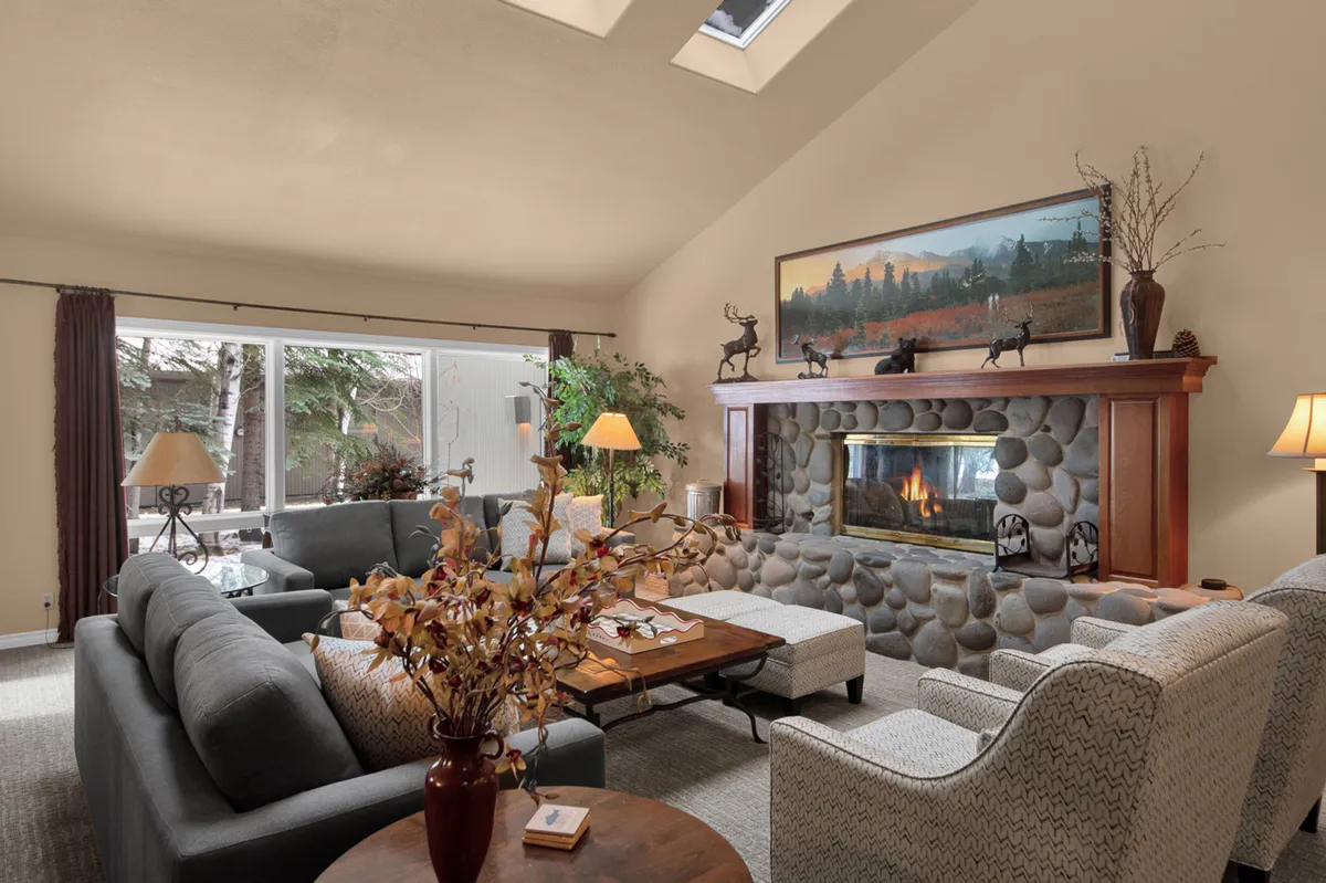 The living area centers around a welcoming fireplace, with comfortable seating arranged to take in both the warmth of the fire and the views through large windows. Light floods the room, and the decor hints at a stylish yet cozy retreat, perfect for gathering during wintertime or enjoying tranquil mornings year-round.