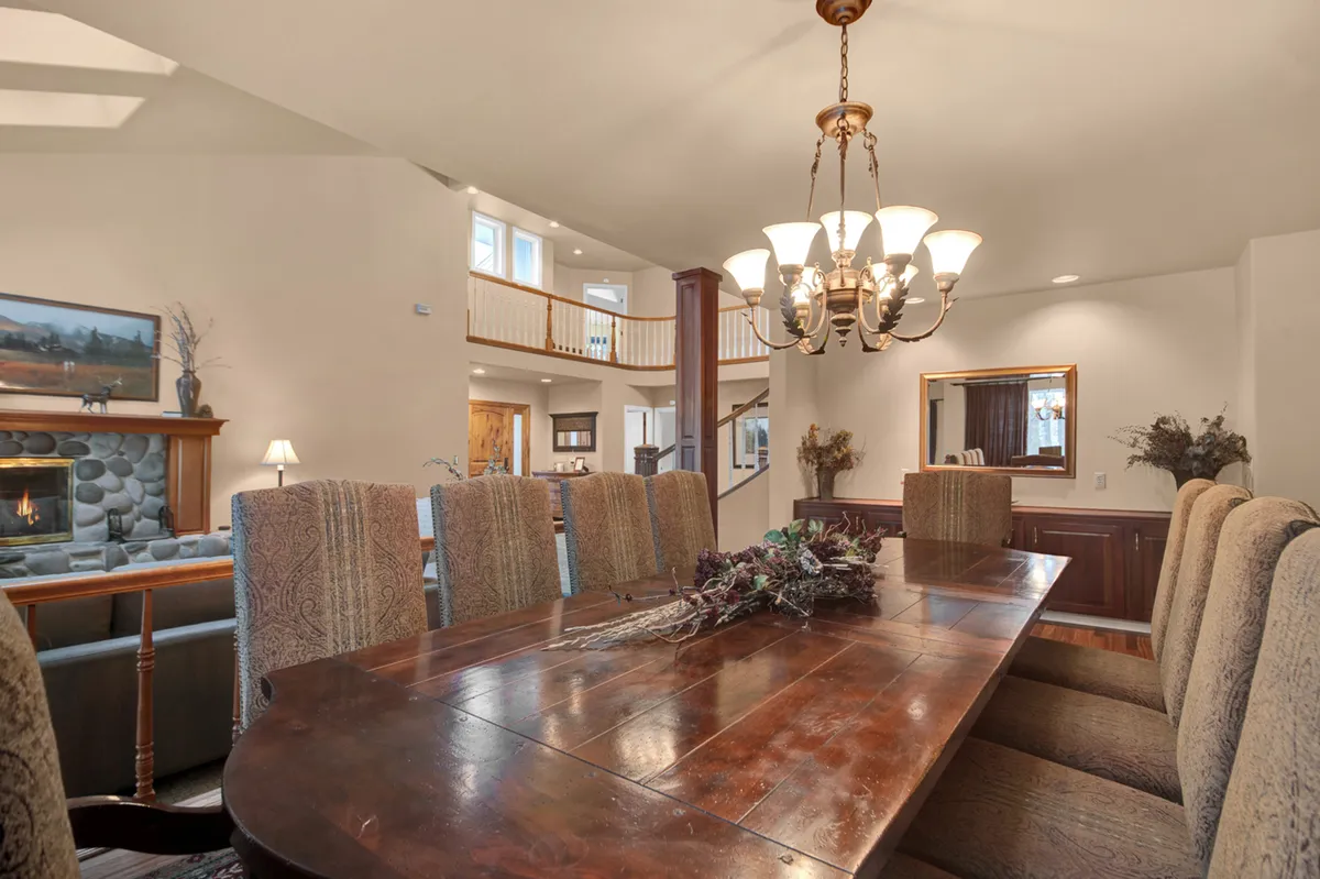 A large dining area with an expansive wooden table, elegant chandelier, and a fireplace in the background. Wooden railings and warm lighting make the space feel both grand and cozy, ideal for group meals and celebrations.