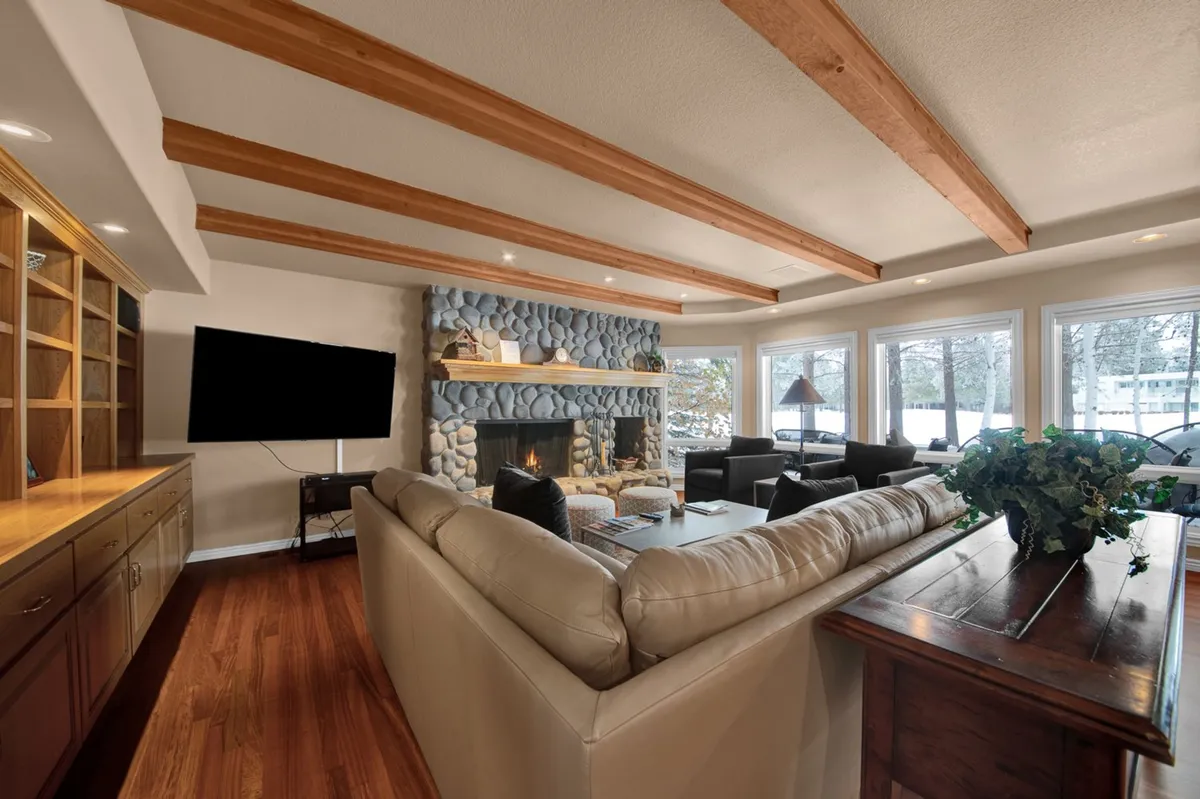 A warm and welcoming living room centered around a striking stone fireplace, complemented by a luxurious leather sofa and exposed wooden beams overhead. Large windows bathe the space in natural light, and the setup is ideal for relaxing, entertaining, or intimate conversations.