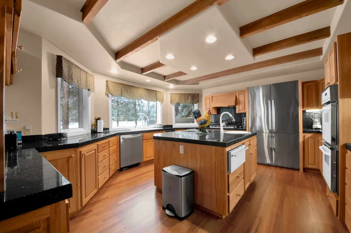 This image shows a welcoming kitchen with a central island and sink, stainless steel appliances, large windows that flood the space with natural light, and wooden beams overhead. The professional fridge and dual cooktops are visible, showcasing the space's culinary capabilities.