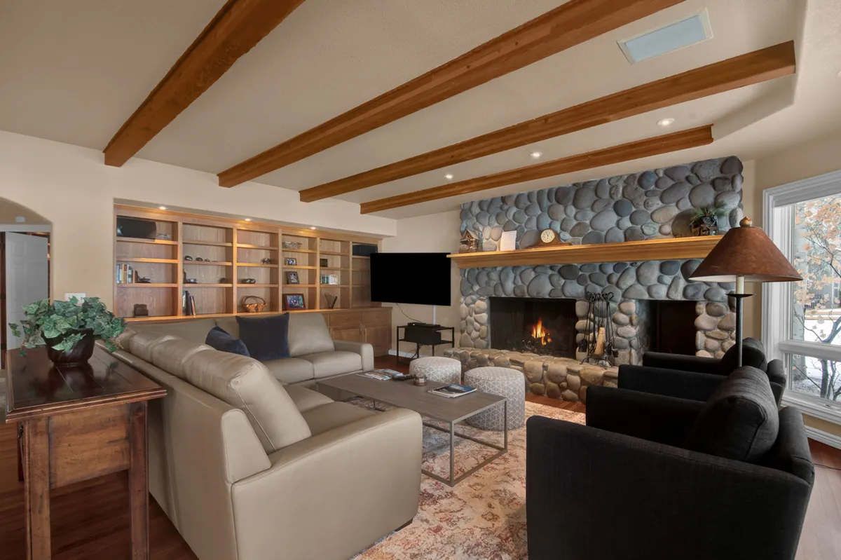 A cozy family room featuring a rustic stone fireplace, wood beams, comfortable seating, and a filled bookshelf. The arrangement is ideal for gathering, reading, watching TV, or enjoying quiet family time.