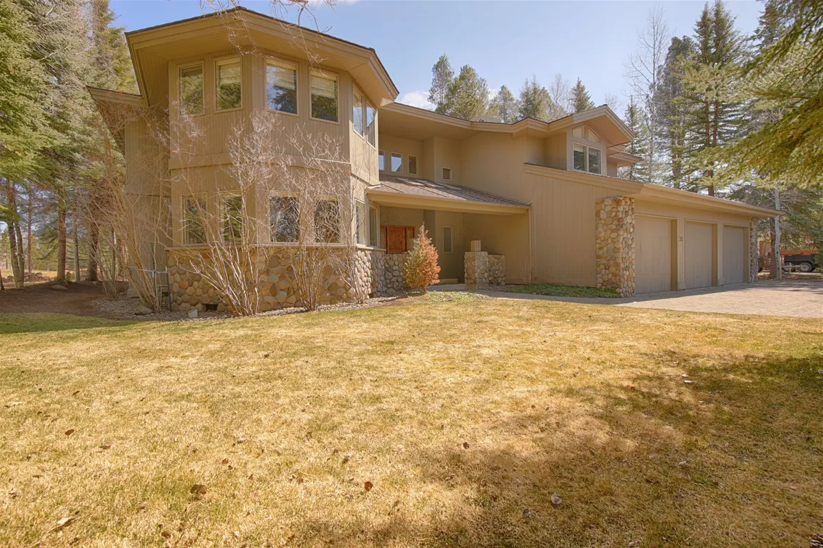 This exterior image showcases the home's unique architectural design set against lush greenery, with prominent stone accents and large windows that offer a glimpse of the inside. The house is surrounded by well-maintained landscaping, emphasizing a seamless connection to the outdoors and the potential for enjoying the surrounding environment.