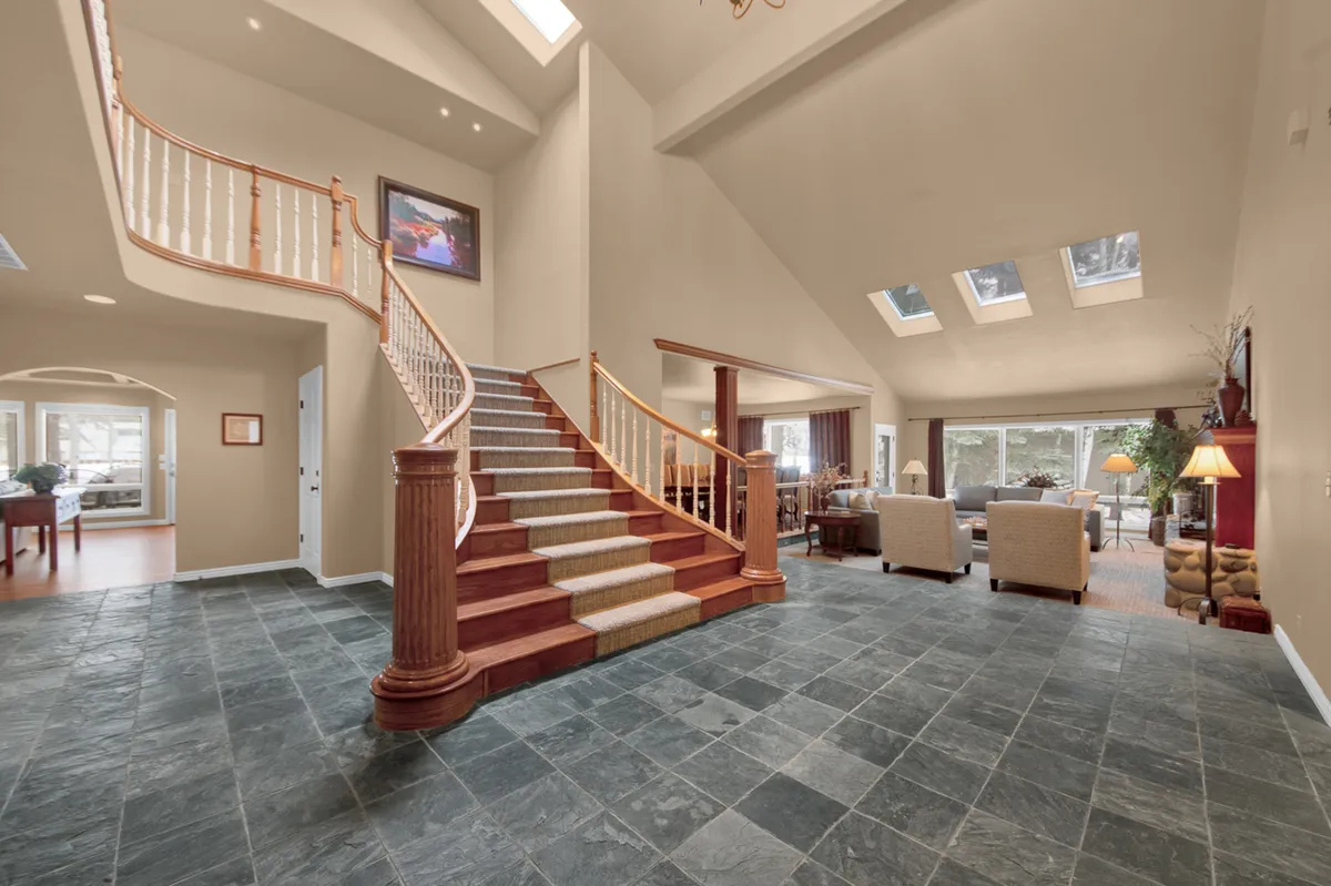 This image showcases a grand staircase rising from a spacious foyer, with large windows that flood the area with natural light. The open living area beyond hints at the welcoming, communal spaces just past the entry. Elegant details and airy design evoke a sense of arrival and comfort.