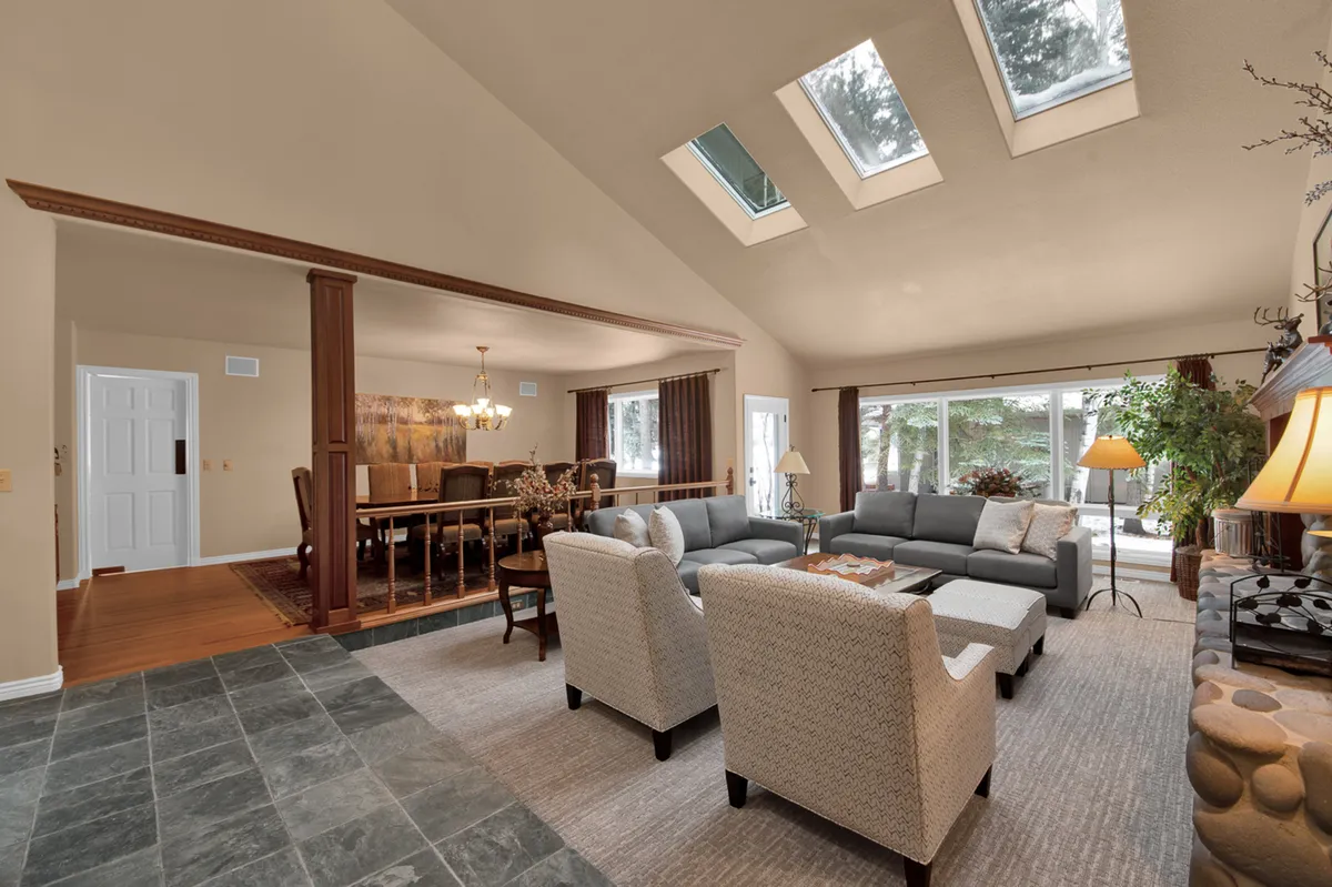A sunlit seating area features a spacious sofa and a dining table, all bathed in natural light from expansive windows that offer a glimpse of outdoor greenery. The scene evokes relaxed summer mornings or snowy afternoons enjoyed from the comfort of home.