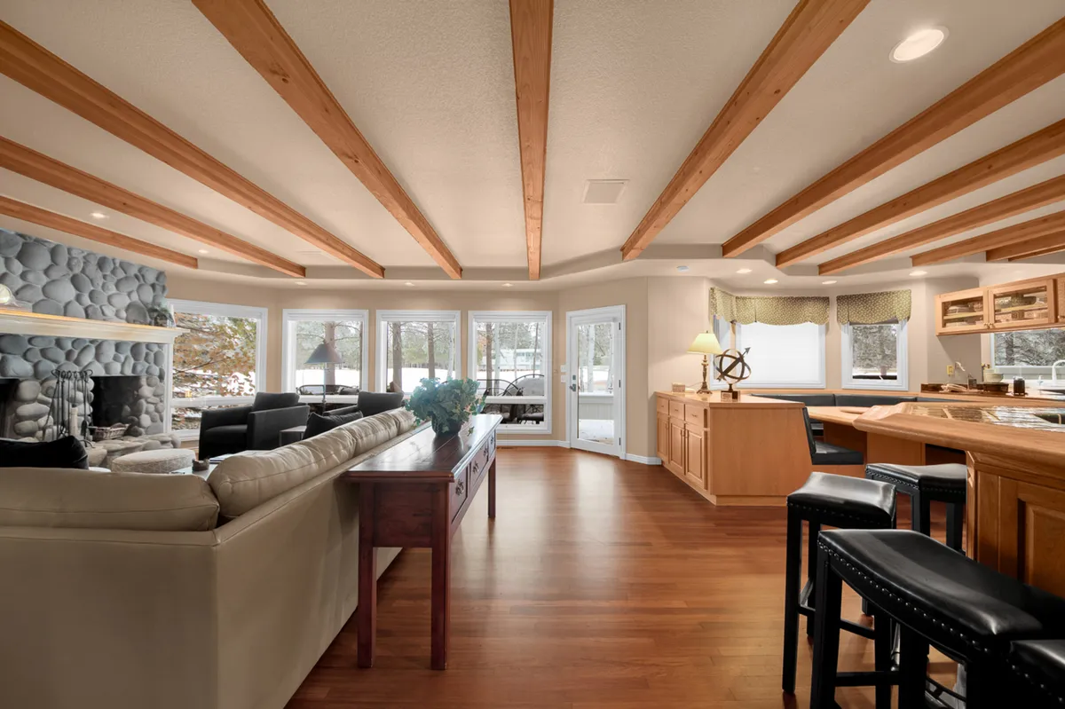 The image captures an open-plan area with a view of the stone fireplace, exposed wooden beams, large windows, and the adjacent kitchen. The inviting layout encourages family gatherings and casual dining, blending the kitchen and social spaces seamlessly.
