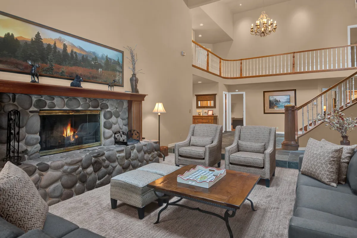 The image showcases a warm, spacious living room centered around a glowing stone fireplace, with elegant sofas arranged for conversation, a striking chandelier overhead, and inviting décor that encourages both relaxation and togetherness. The space radiates coziness, perfect for family gatherings or unwinding after a day of adventure.