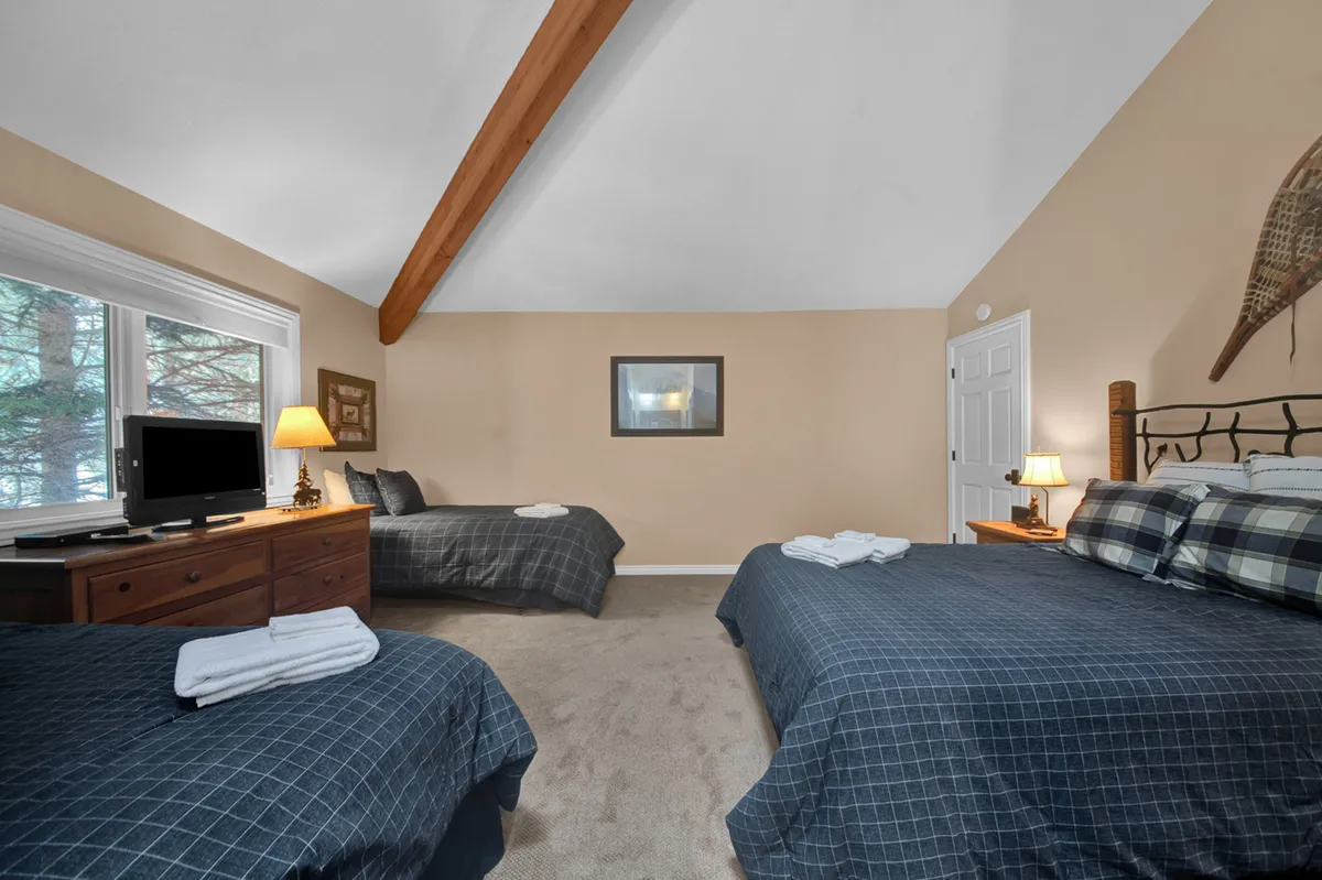 A spacious bedroom with two large beds, a wooden dresser, television, lamp, and decorative wall hanging. The room exudes a comfortable and welcoming mood, ideal for families or groups sharing a space.